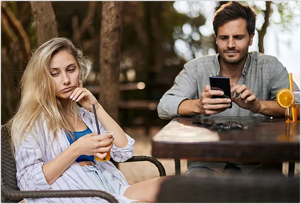Ein Pärchen sitzt mit Drinks an einem Tisch im Freien, er ist am Handy, sie schaut gelangweilt drein.